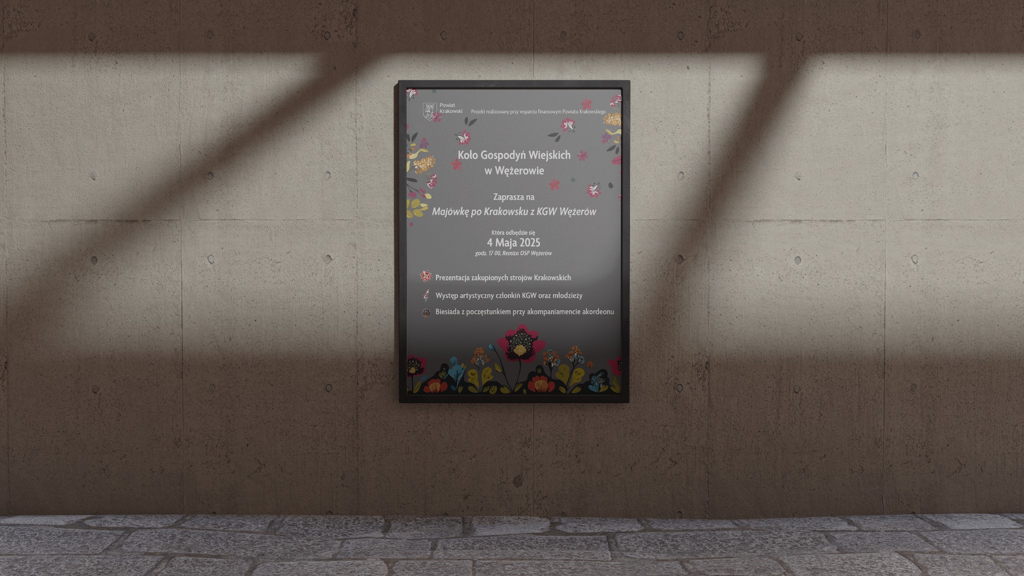 Plakat promujący Majówkę po Krakowsku z KGW Wężerów, z motywami folklorystycznymi. Grafika w ramie wisi na betonowej ścianie, oświetlona z góry. Kolorystyka stonowana, z akcentami kwiatowymi.