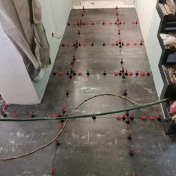 R.S Wykonczenia - Układanie płytek podłogowych z użyciem systemu poziomowania w Bytomiu. Widoczne płytki w odcieniach szarości i czerwone kliny dystansowe. Przewody na podłodze.