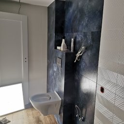 R.S Wykonczenia - Nowoczesna łazienka z bidetem na tle ściany z ciemnych płytek imitujących kamień i jasnych płytek z wzorem chevron. Widoczne akcesoria na półce.