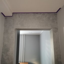 R.S Wykonczenia - Ściana z efektem dekoracyjnym, zabezpieczona taśmą malarską przy suficie, widok z perspektywy niskiego kąta, wnętrze w szarościach.