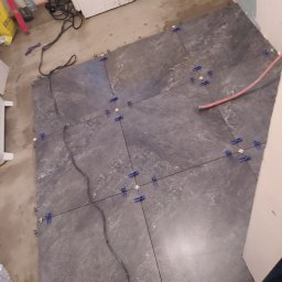 R.S Wykonczenia - Układanie ciemnych płytek podłogowych z niebieskimi krzyżykami dystansowymi. Widoczne przewody i narzędzia. Fragment ściany w tle. Praca w toku.