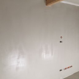 R.S Wykonczenia - Ściana po szpachlowaniu i malowaniu na jasny odcień. Widoczne puszki elektryczne i otwory pod gniazdka. Belka stropowa w rogu sufitu.