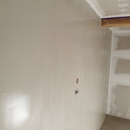 R.S Wykonczenia - Gładka, jasna ściana po szpachlowaniu z widocznymi puszkami elektrycznymi, drewnianymi elementami konstrukcyjnymi sufitu i fragmentem regału z płyty KG w rogu pokoju.