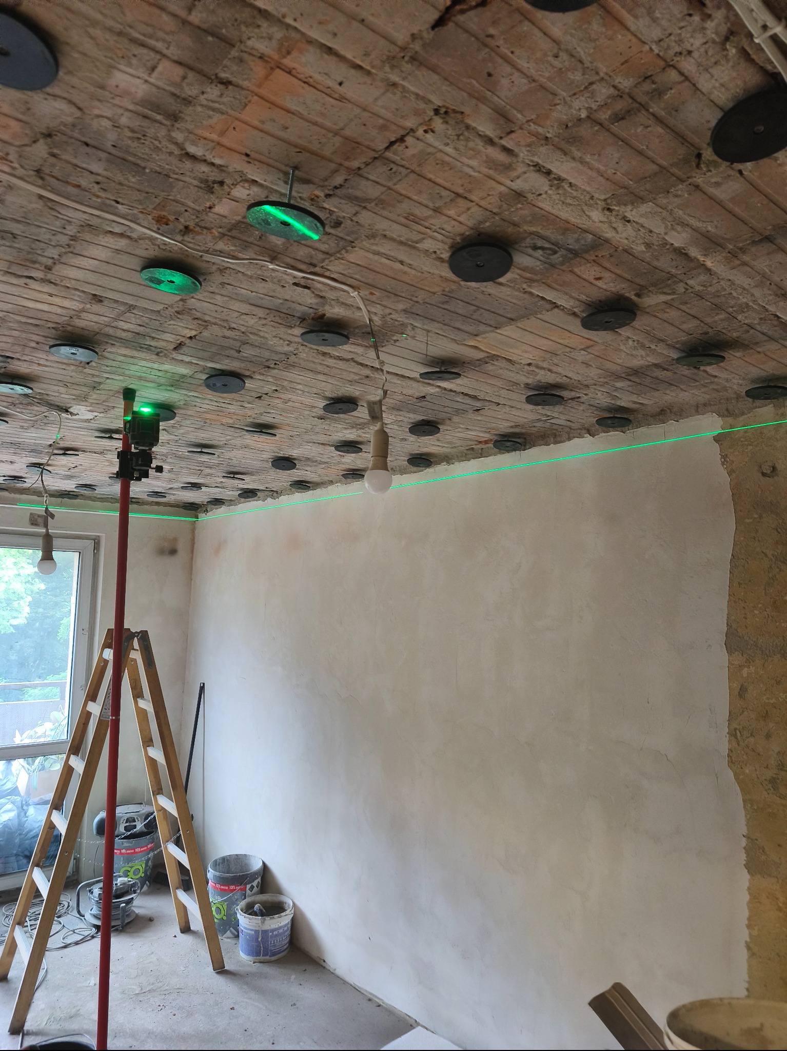 Pokój w trakcie remontu: ściany po szpachlowaniu, laser krzyżowy wyznacza poziom, widoczny sufit z przymocowanymi elementami montażowymi.