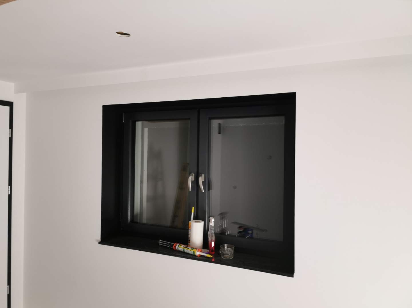 Białe ściany, czarna rama okienna z akcesoriami na parapecie. Widoczne świeżo pomalowane powierzchnie i wykończone detale.