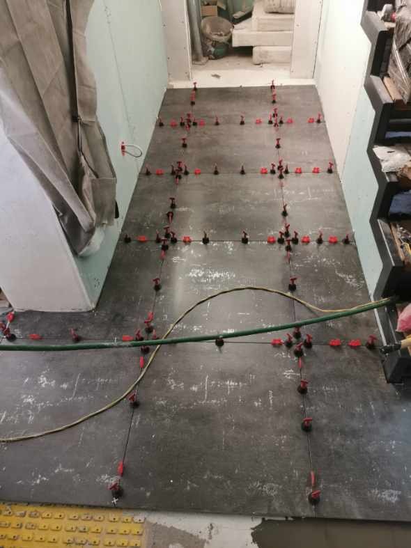 Układanie płytek podłogowych z użyciem systemu poziomowania w Bytomiu. Widoczne płytki w odcieniach szarości i czerwone kliny dystansowe. Przewody na podłodze.