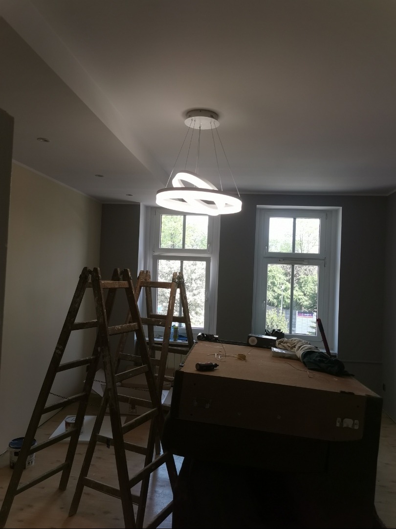 Pokój w trakcie malowania z dwoma oknami, drabiny malarskie i nowoczesna lampa wisząca. Widoczne przygotowania do malowania ścian na szaro.