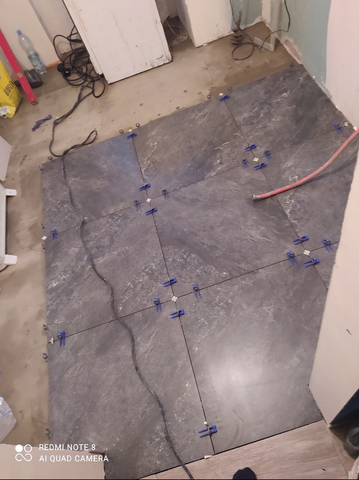 Układanie ciemnych płytek podłogowych z niebieskimi krzyżykami dystansowymi. Widoczne przewody i narzędzia. Fragment ściany w tle. Praca w toku.