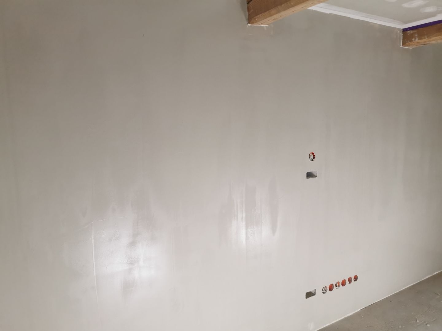 Ściana po szpachlowaniu i malowaniu na jasny odcień. Widoczne puszki elektryczne i otwory pod gniazdka. Belka stropowa w rogu sufitu.