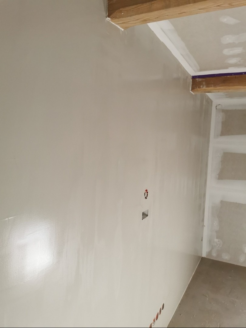 Gładka, jasna ściana po szpachlowaniu z widocznymi puszkami elektrycznymi, drewnianymi elementami konstrukcyjnymi sufitu i fragmentem regału z płyty KG w rogu pokoju.