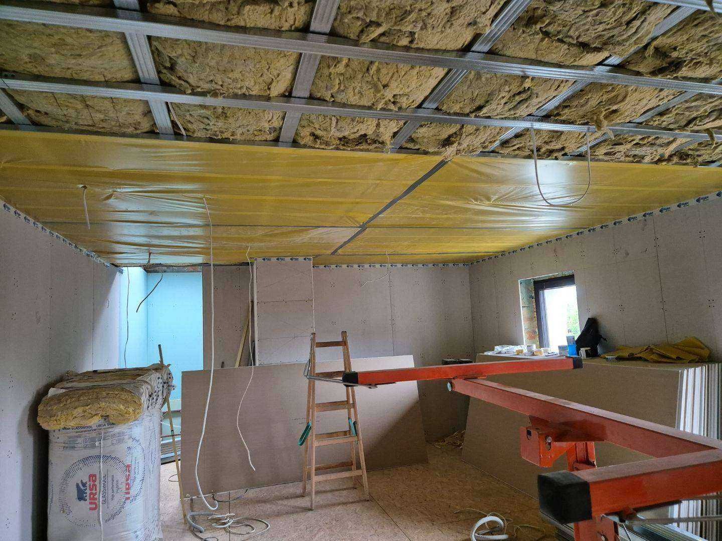 Montaż izolacji i płyt gipsowych na suficie podwieszanym. Widoczne profile, wełna mineralna, folia paroizolacyjna i częściowo zamontowane płyty.