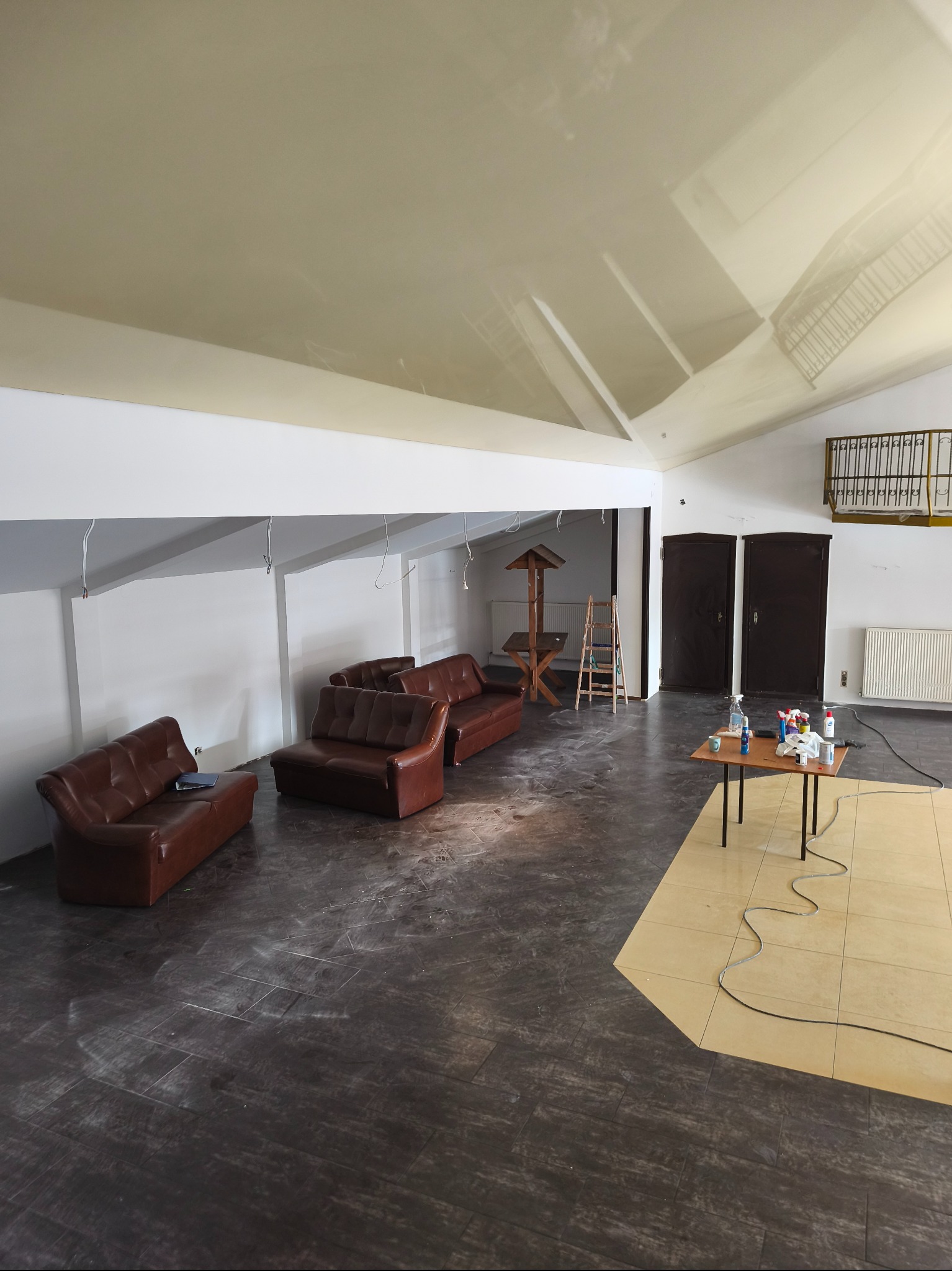 Przestronne wnętrze w trakcie wykańczania: brązowe sofy, dwukolorowa podłoga (ciemne panele i jasne płytki), białe ściany i wysoki sufit. Widoczne narzędzia i drabina.