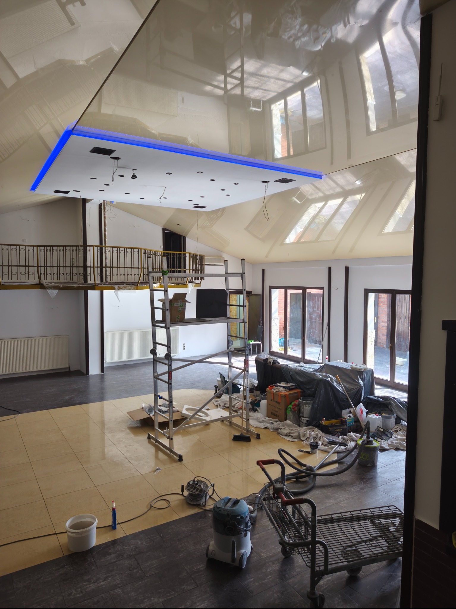 Duże pomieszczenie w trakcie prac wykończeniowych, z widocznym rusztowaniem, narzędziami i sufitem podwieszanym z niebieskim oświetleniem LED.