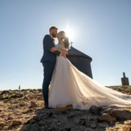Para młoda w objęciach na tle g&oacute;rskiego krajobrazu z charakterystycznym budynkiem. Słońce dodaje blasku, a długa suknia ślubna opada na kamieniste podłoże.