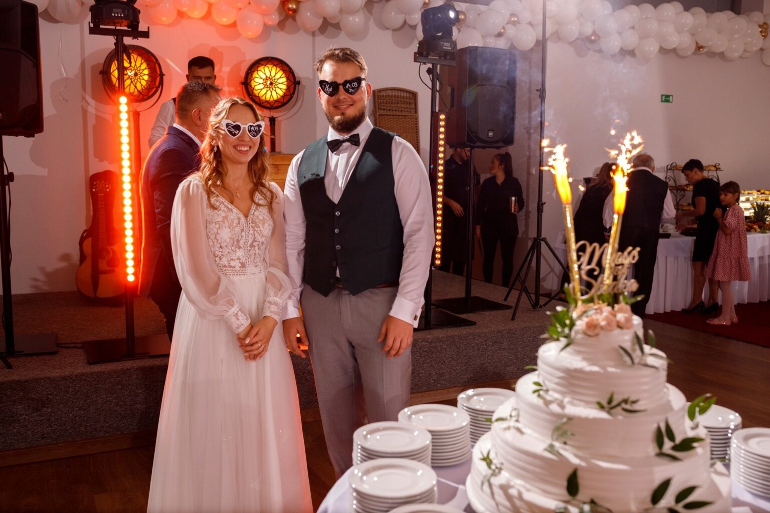 Uśmiechnięta para młoda w okularach w kształcie serc, z tortem weselnym ze sztucznymi ogniami, na tle sali z dekoracjami i gośćmi.