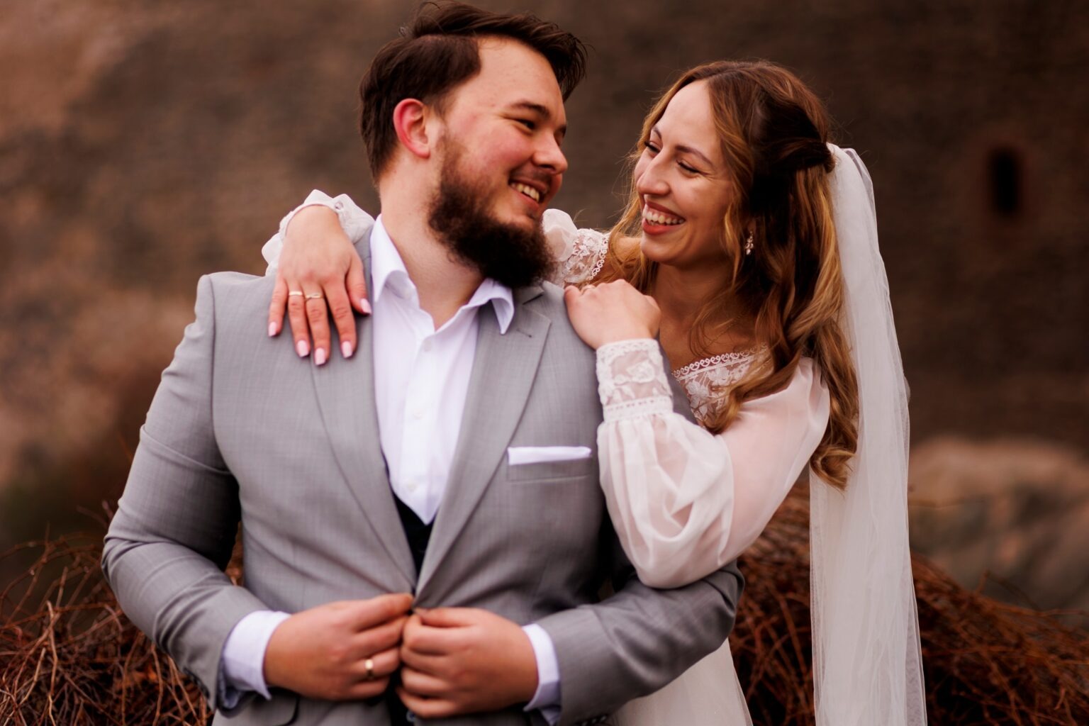 Uśmiechnięta para młoda w objęciach na tle ceglanej ściany. Panna młoda w sukni z koronkowymi rękawami, pan młody w szarym garniturze. Widoczne obrączki.