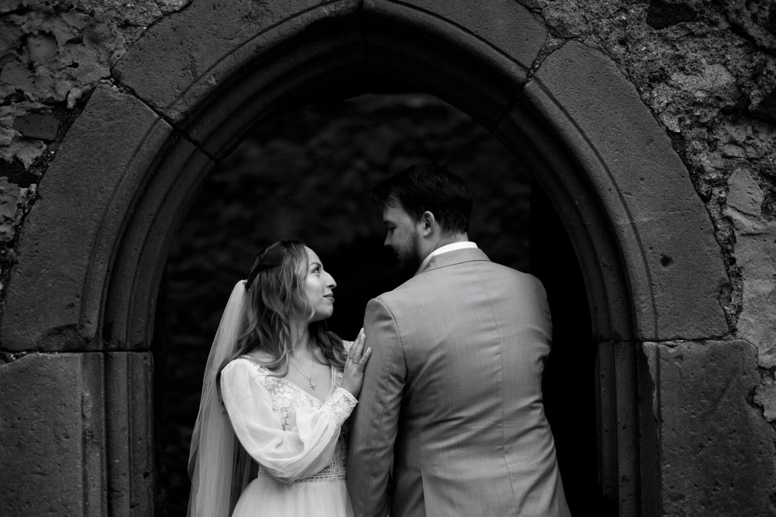 Czarno-biała fotografia ślubna z Legnicy: para młoda patrzy na siebie, stojąc w kamiennym łuku. Panna młoda w sukni z koronką, pan młody w garniturze.