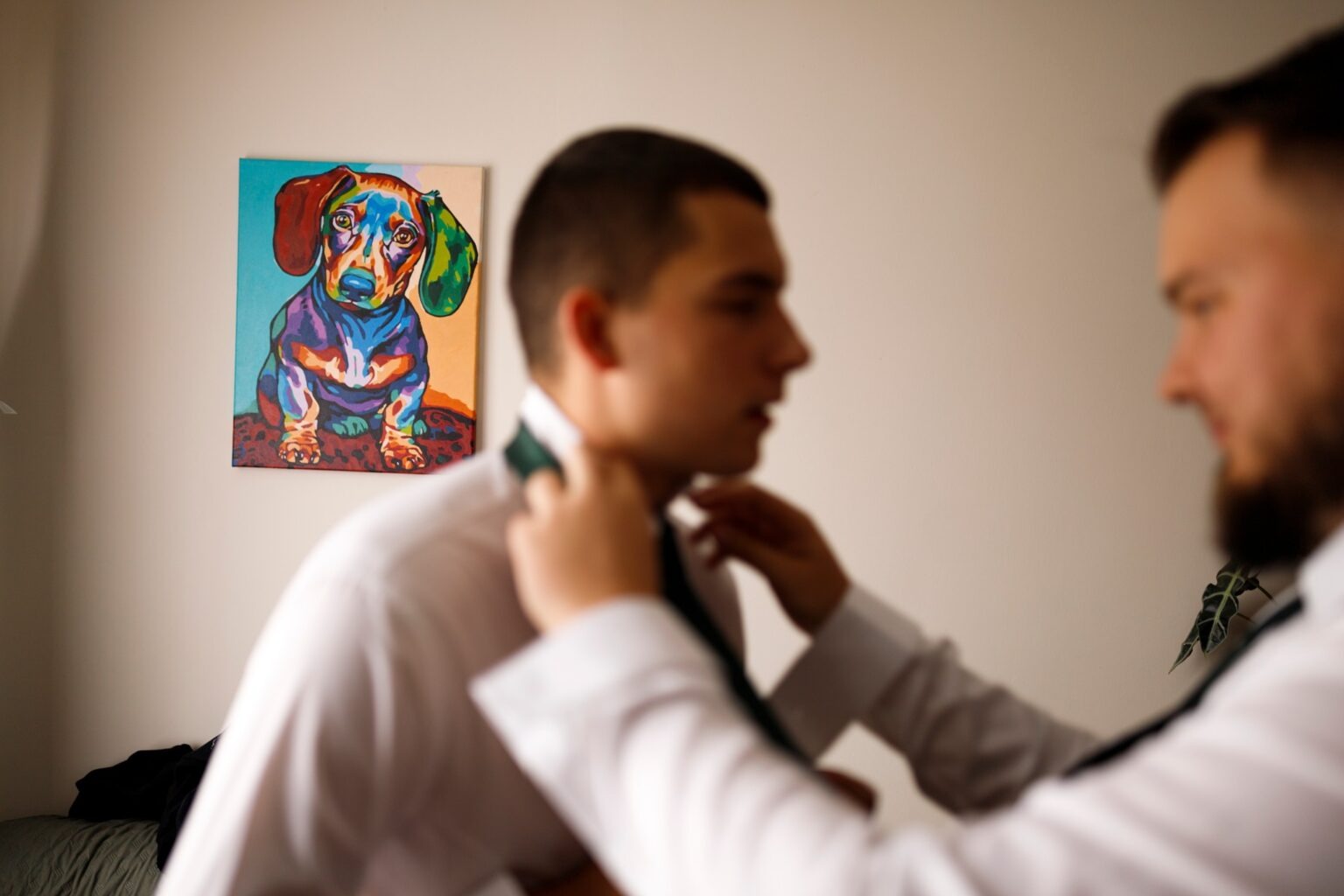 Mężczyzna poprawia krawat drugiemu mężczyźnie. W tle na ścianie wisi kolorowy obrazek przedstawiający psa rasy jamnik.