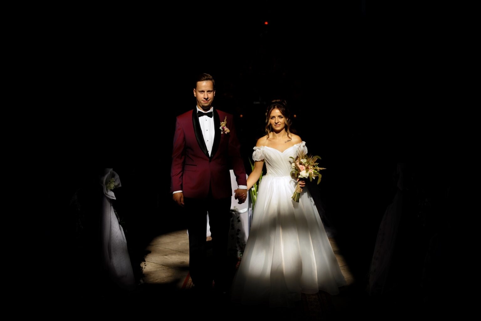 Para młoda, on w bordowym smokingu, ona w białej sukni, idą oświetloną alejką w ciemnym wnętrzu, trzymając się za ręce. Ujęcie w głębokim kontraście.
