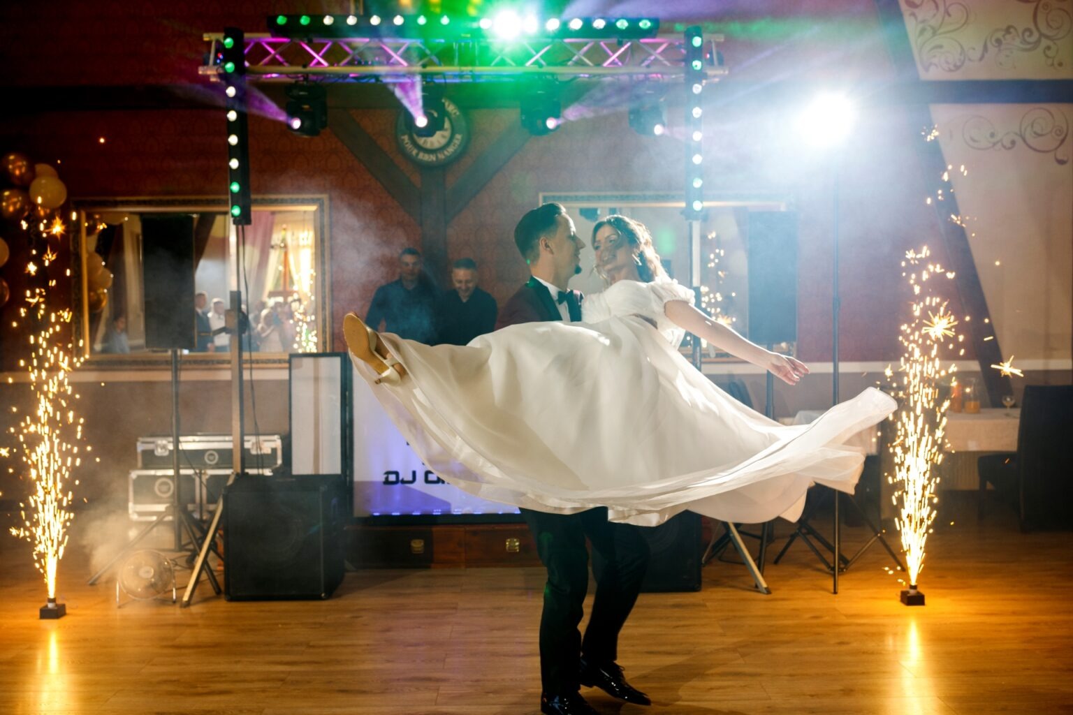 Pan młody trzyma pannę młodą na rękach podczas pierwszego tańca, w tle efekty pirotechniczne, dekoracje i oświetlenie sali weselnej.