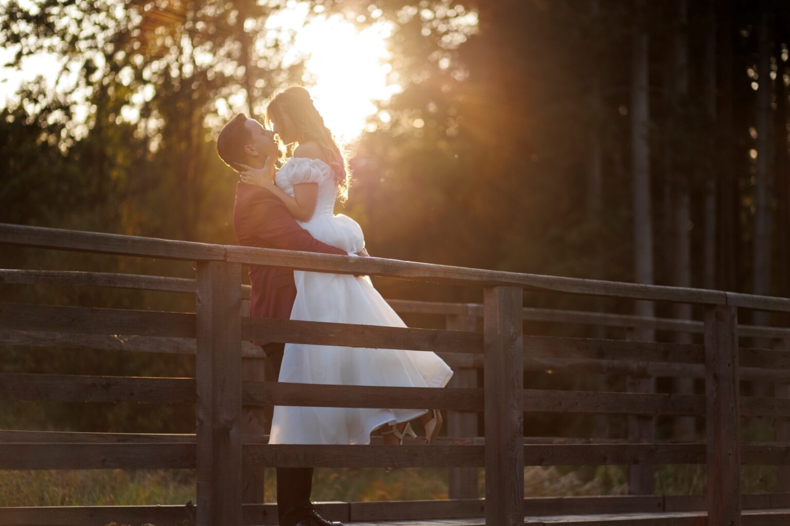 Para młoda na drewnianym mostku w lesie, on trzyma ją w ramionach, ciepłe światło zachodzącego słońca tworzy romantyczną atmosferę.