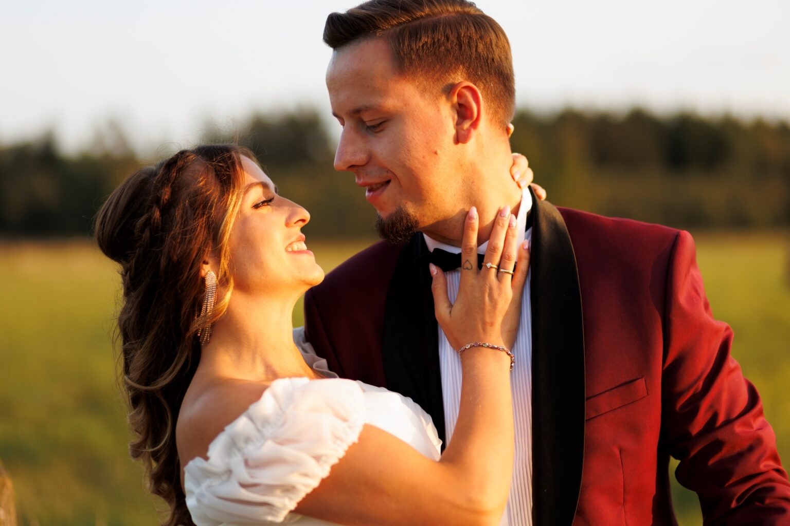 Uśmiechnięta para w strojach ślubnych, spoglądająca na siebie w plenerze, z widoczną biżuterią i tatuażem serca na dłoni kobiety. Ciepłe, naturalne światło.