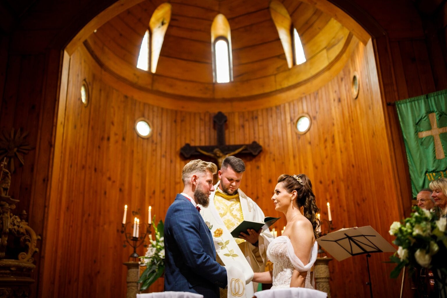 Para młoda podczas ceremonii ślubnej w drewnianym kościele z oryginalnymi oknami. Ksiądz czyta przysięgę małżeńską. Świece i kwiaty.