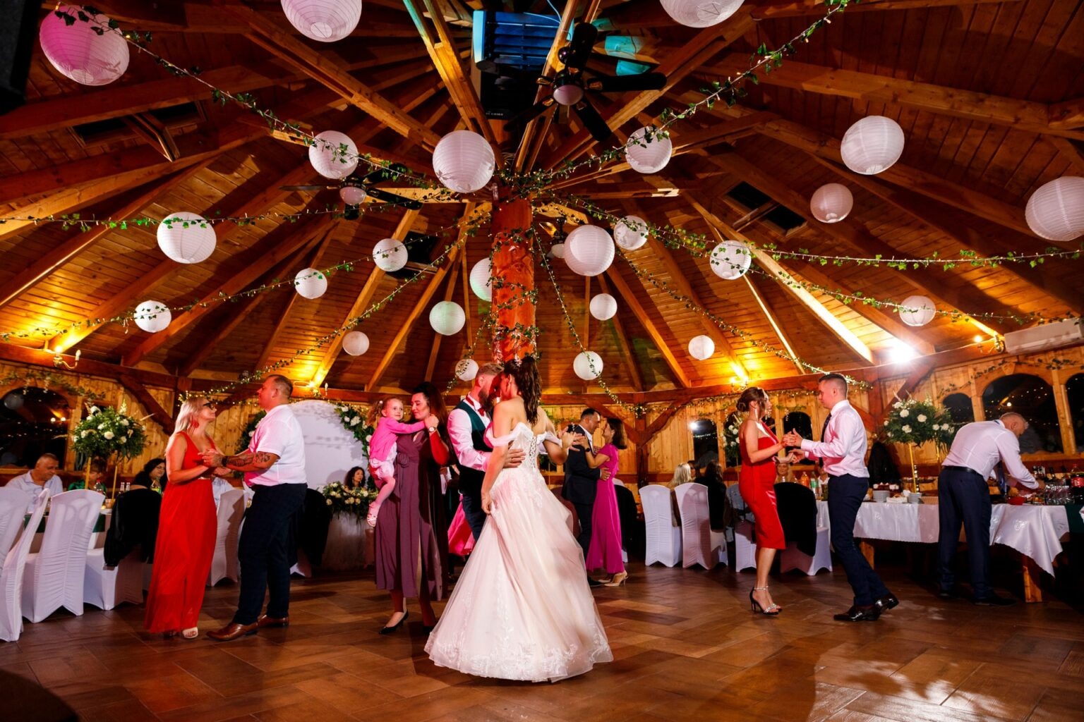 Para młoda tańczy na parkiecie w drewnianej sali weselnej, udekorowanej lampionami i girlandami, w otoczeniu gości.