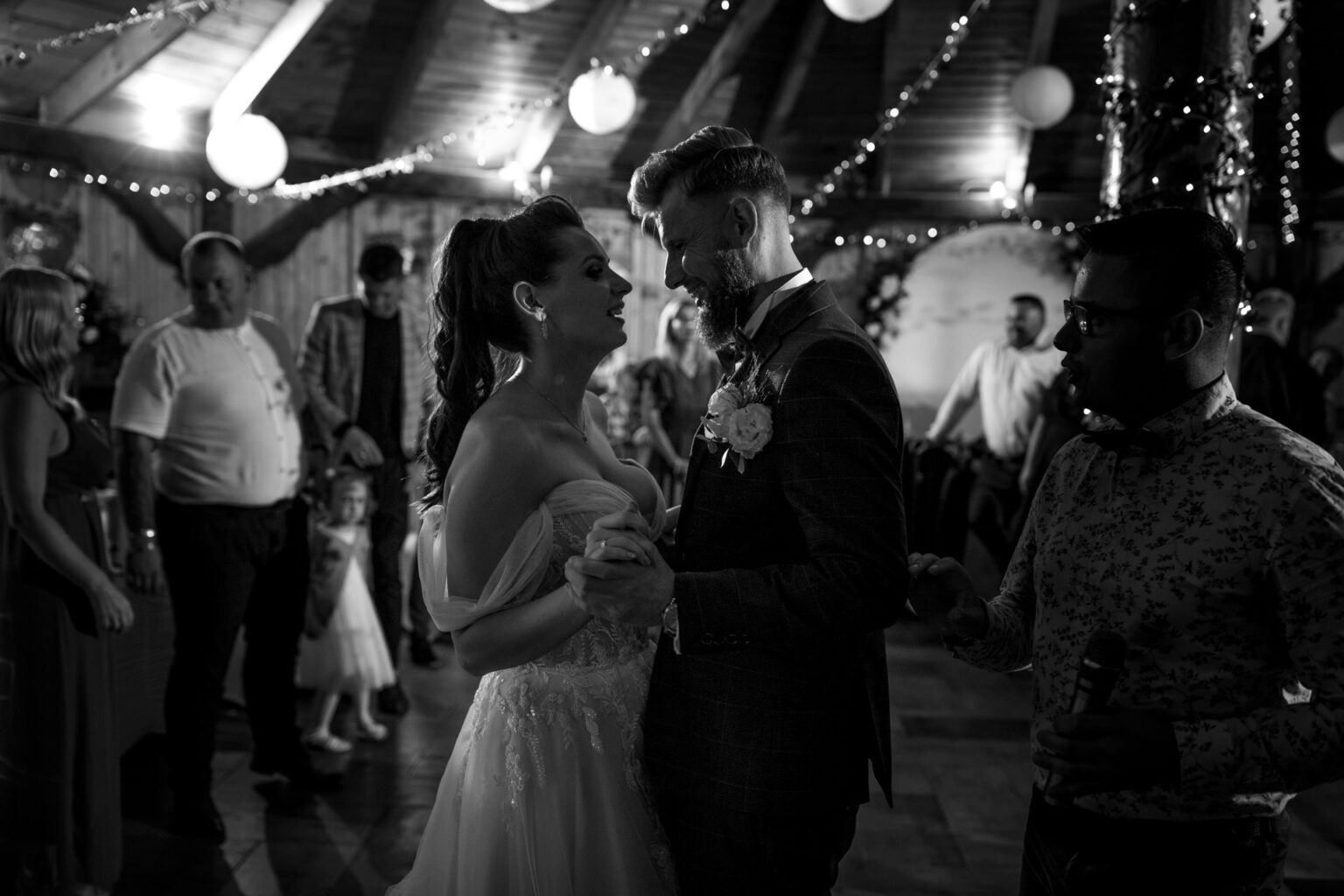 Czarno-białe zdjęcie pary młodej w tańcu podczas wesela w rustykalnej sali, z gośćmi i dekoracjami świetlnymi w tle. Pan młody w ciemnym garniturze, panna młoda w sukni.