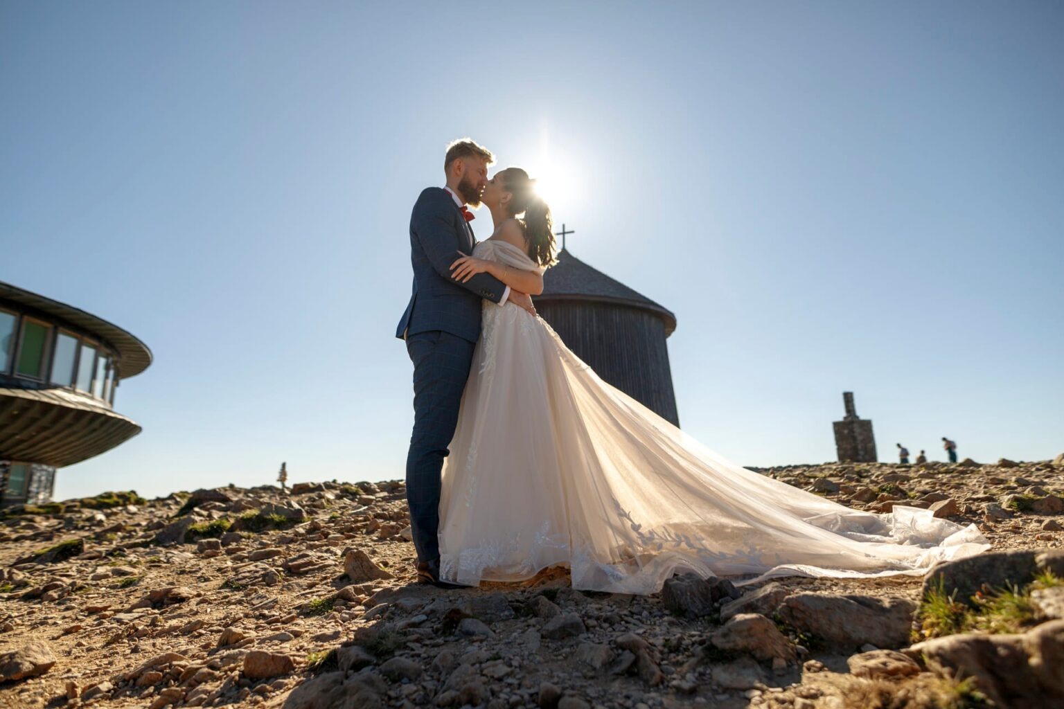 Para młoda w objęciach na tle górskiego krajobrazu z charakterystycznym budynkiem. Słońce dodaje blasku, a długa suknia ślubna opada na kamieniste podłoże.