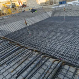 Siatka zbrojeniowa ułożona na betonowej płycie fundamentowej, widoczne wystające pręty i w tle wykop z maszynami budowlanymi.