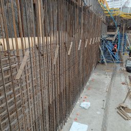 Patryk Opara P.O.Z. - Zbrojenie pionowej ściany wykopu zabezpieczonej ścianką berlińską, widoczne elementy szalunku i pracownik w kasku w tle.