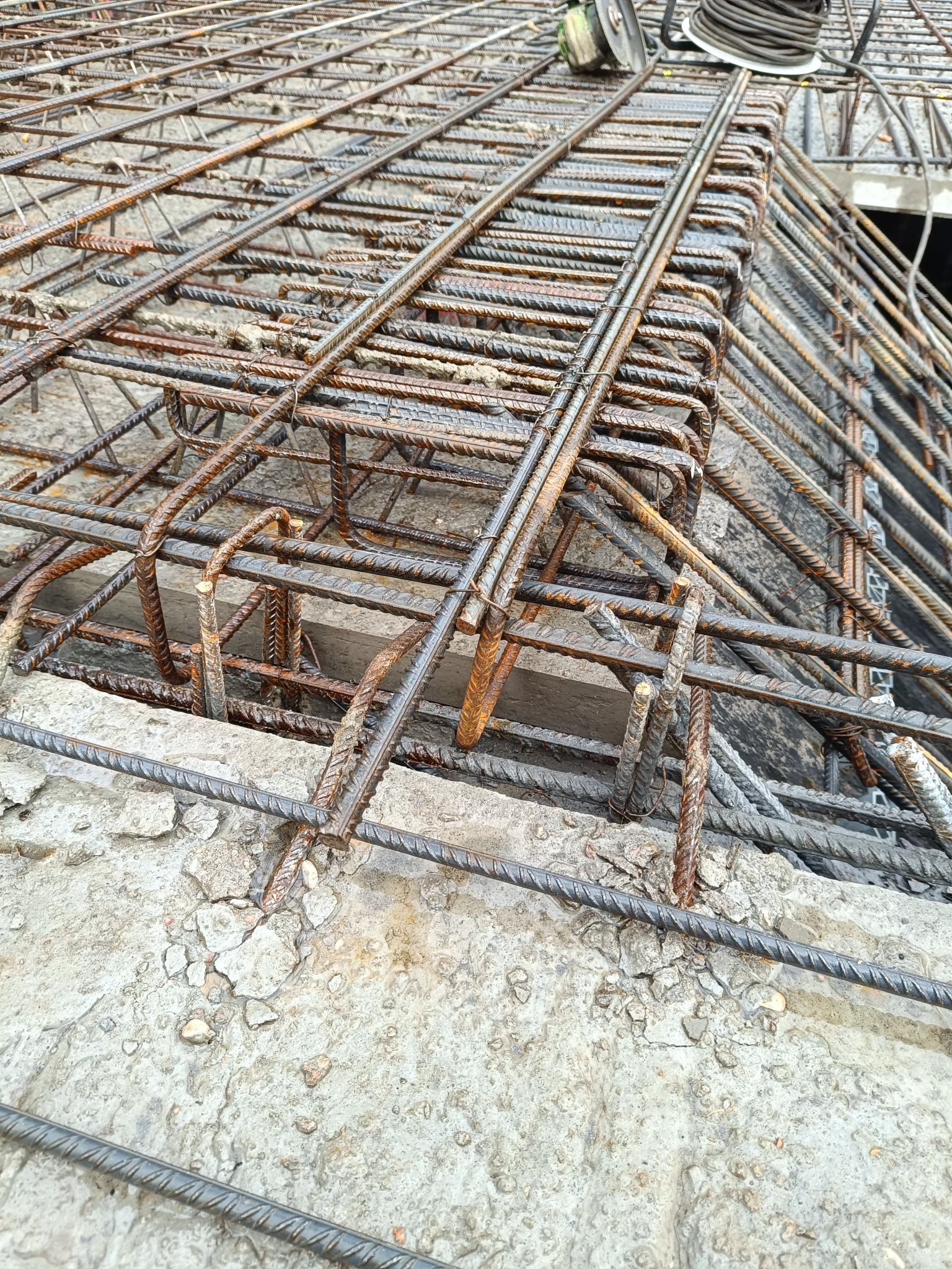 Siatka zbrojeniowa ułożona na szalunku fundamentów, widoczne fragmenty betonu i wystające pręty, z góry widoczny sprzęt budowlany.