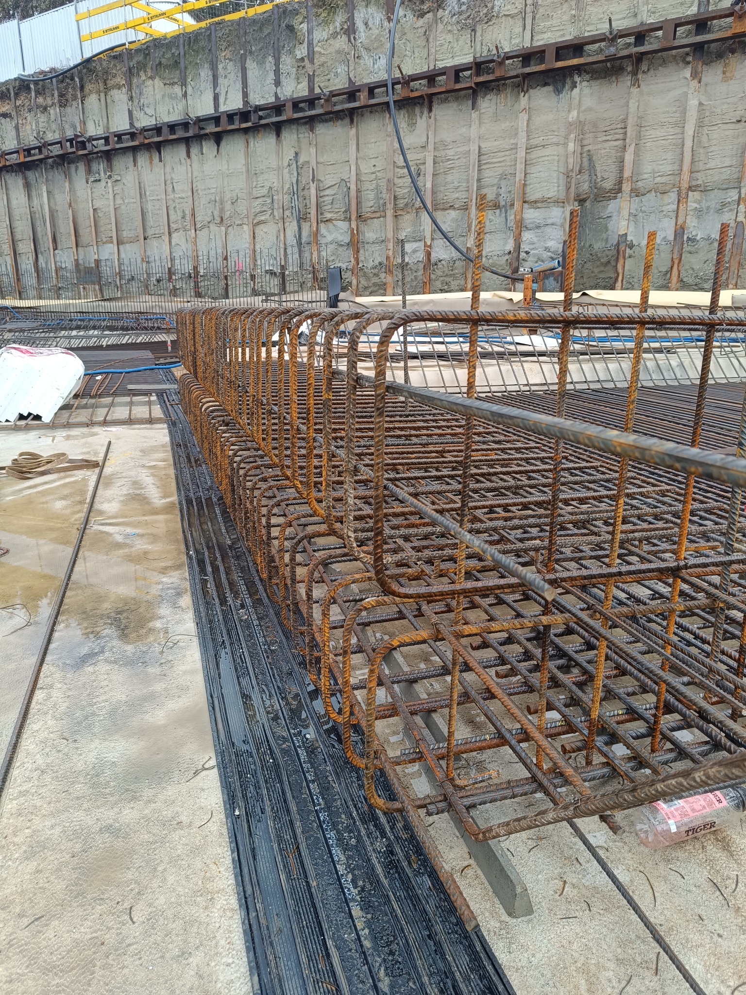 Szkielet zbrojenia fundamentów z widocznym deskowaniem ziemnym w tle, przygotowany do zalania betonem, z butelką wody mineralnej obok.