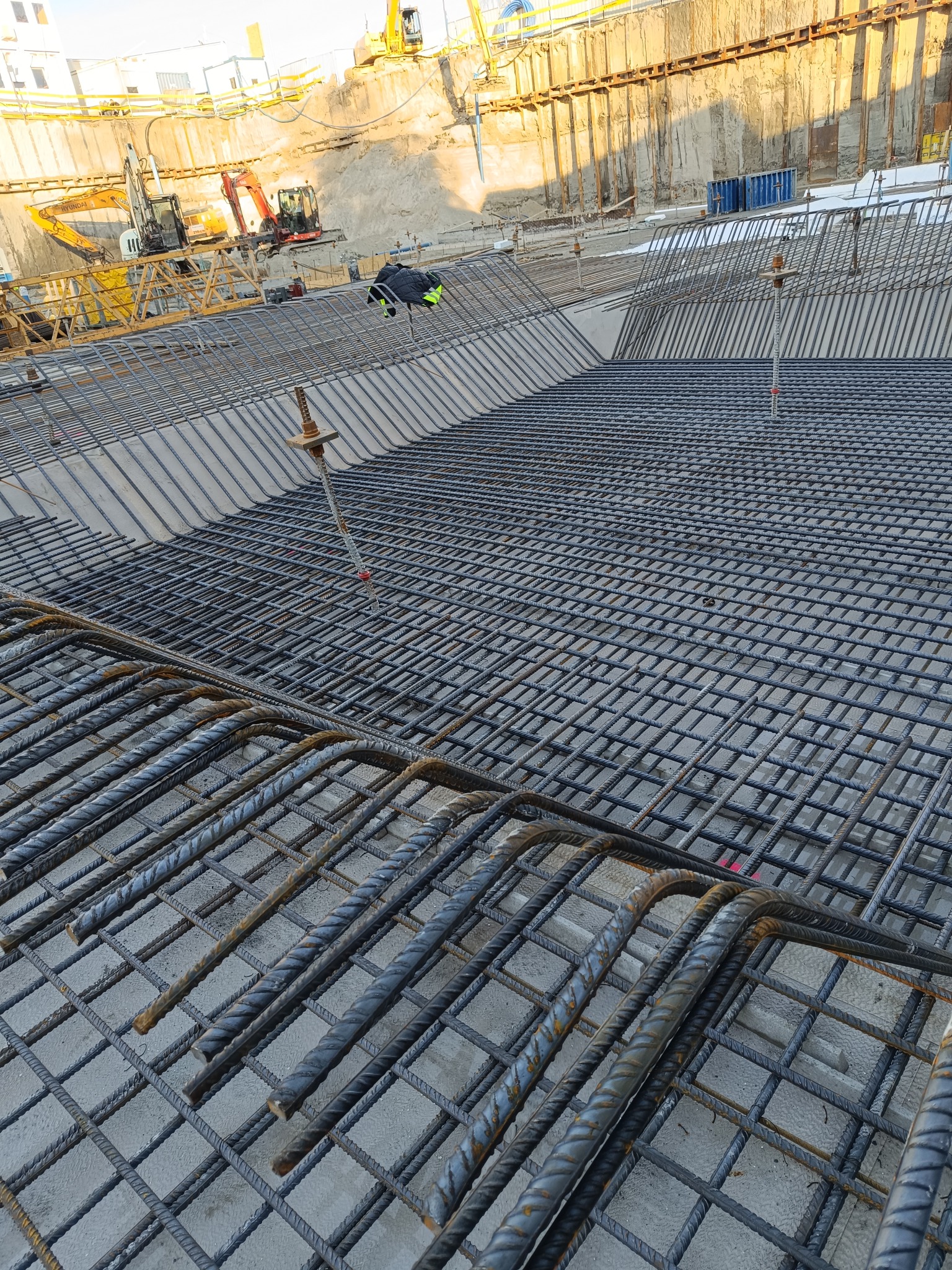 Siatka zbrojeniowa ułożona na betonowej płycie fundamentowej, widoczne wystające pręty i w tle wykop z maszynami budowlanymi.