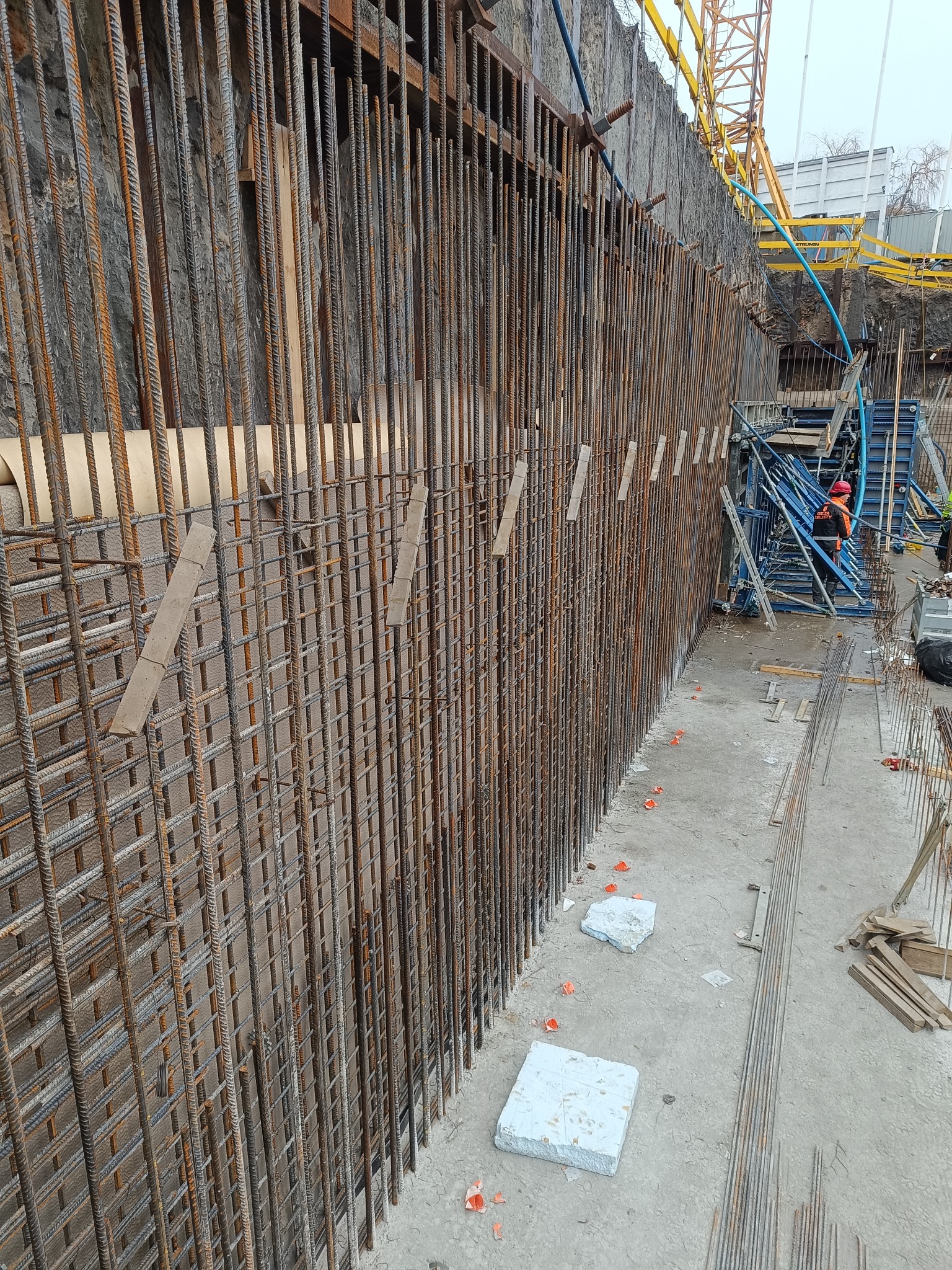 Zbrojenie pionowej ściany wykopu zabezpieczonej ścianką berlińską, widoczne elementy szalunku i pracownik w kasku w tle.