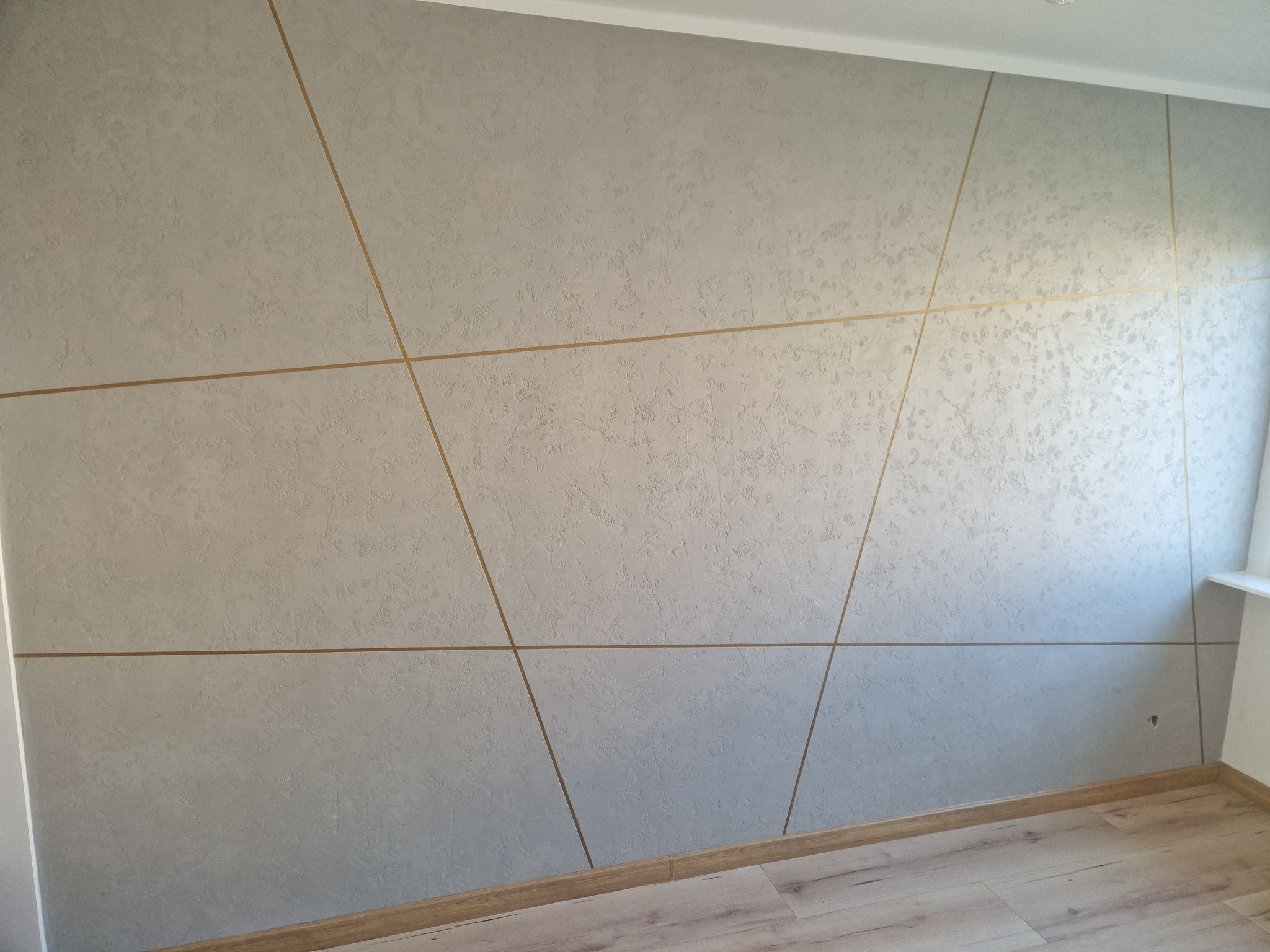 Ściana z dekoracyjną, szarą fakturą, podzielona złotymi listwami na geometryczne pola, widoczny fragment podłogi z paneli.