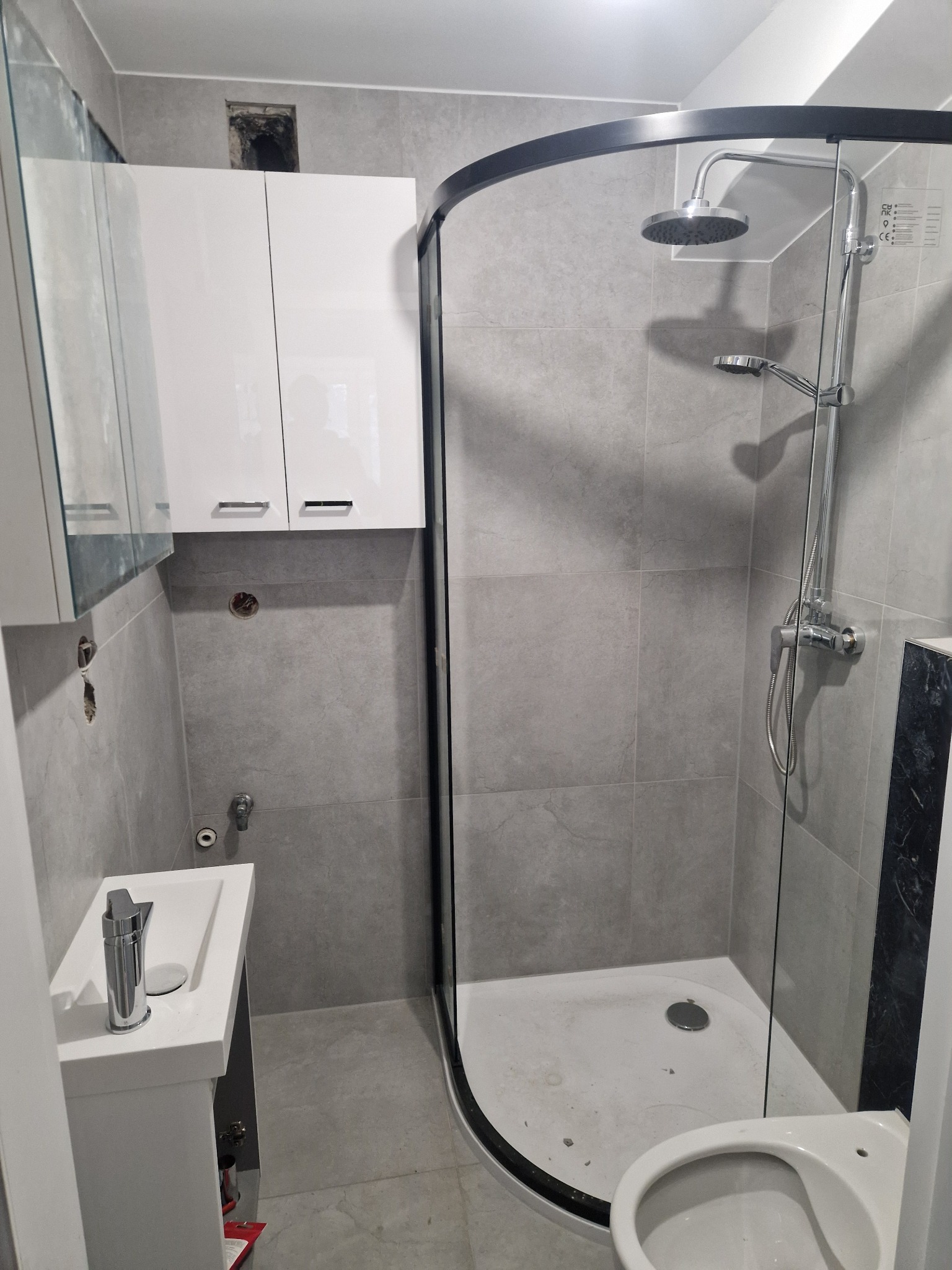 Ukończona łazienka z szarymi płytkami, kabiną prysznicową z czarną ramą, białą szafką nad umywalką i toaletą, widoczne niedokończone otwory w ścianie.