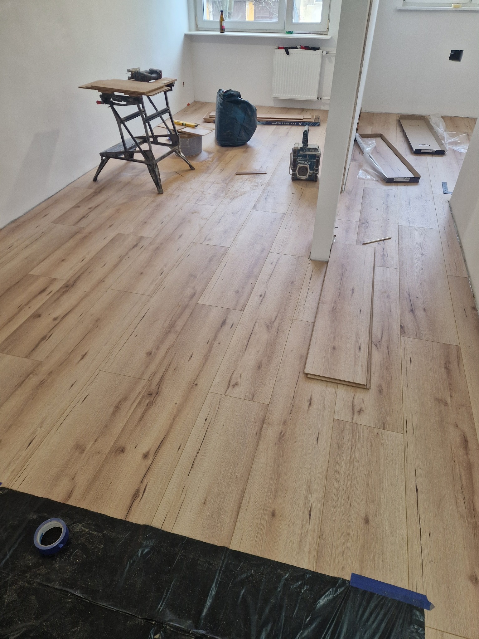 Układanie paneli podłogowych z widocznymi narzędziami: skrzynką narzędziową Makita, stołem roboczym, poziomnicą oraz nieużywanymi panelami w opakowaniach.