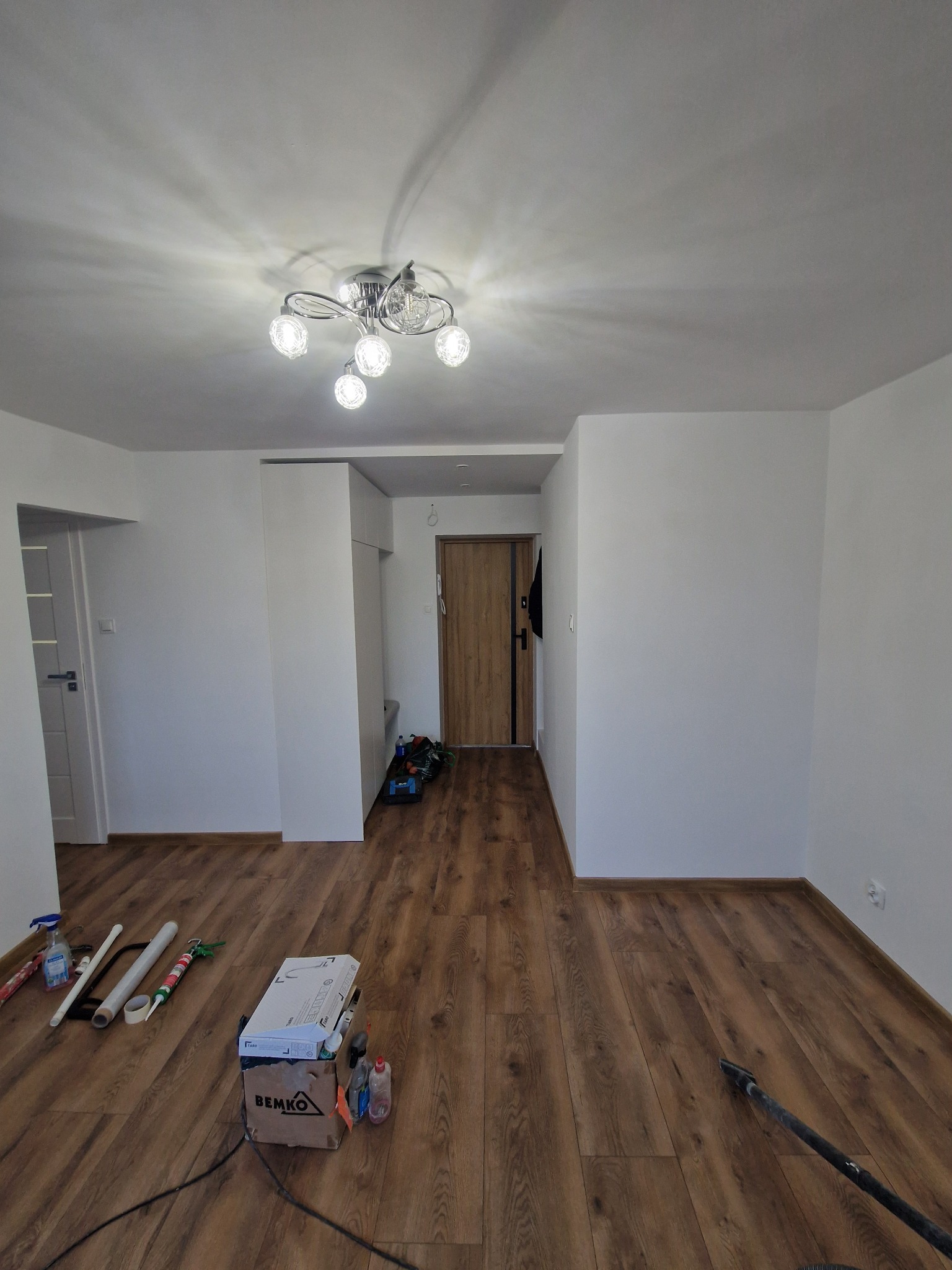 Wnętrze mieszkania po odmalowaniu ścian na biało, z drewnianą podłogą, widoczne narzędzia i materiały malarskie przy ścianie, oraz lampa sufitowa z sześcioma żarówkami.