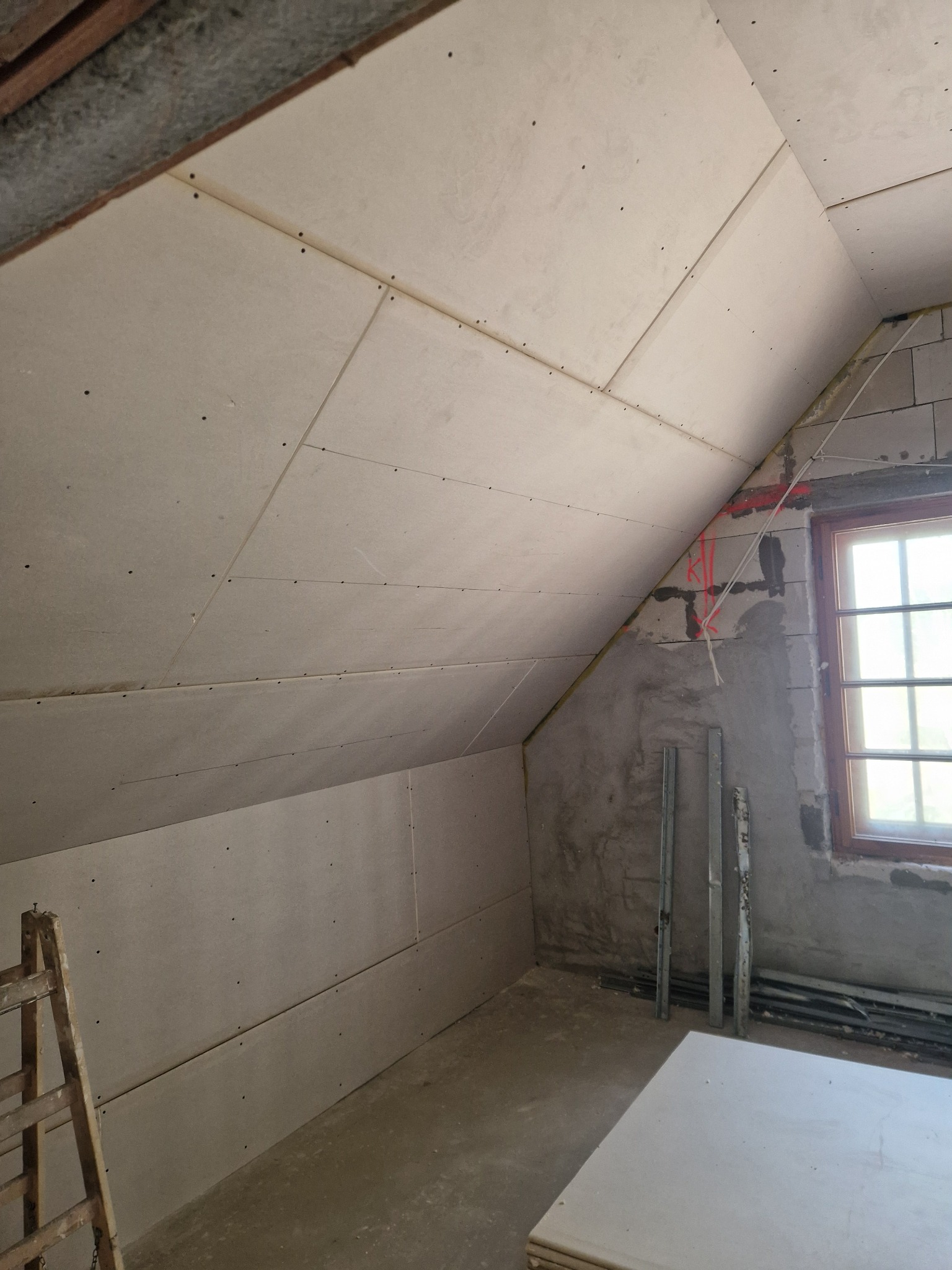 Poddasze w trakcie montażu płyt gipsowo-kartonowych na skosach i ścianach, widoczne wkręty, fragment okna i drabina oparta o ścianę, surowy stan wykończenia.