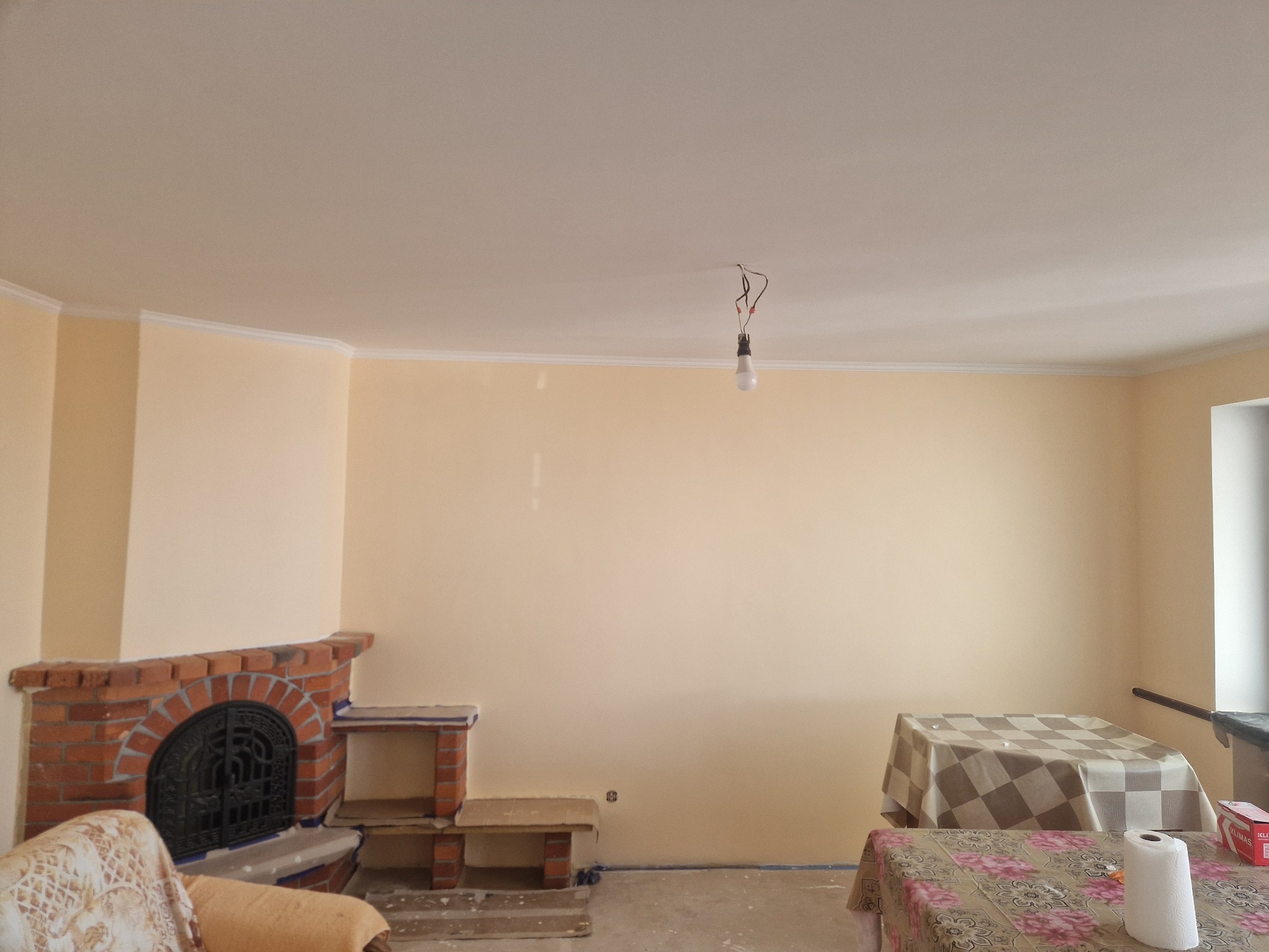 Wnętrze pokoju w trakcie malowania ścian na jasny beż, z widocznym ceglanym kominkiem z żeliwnym wkładem, niezabezpieczonymi meblami i wiszącą żarówką na kablu.