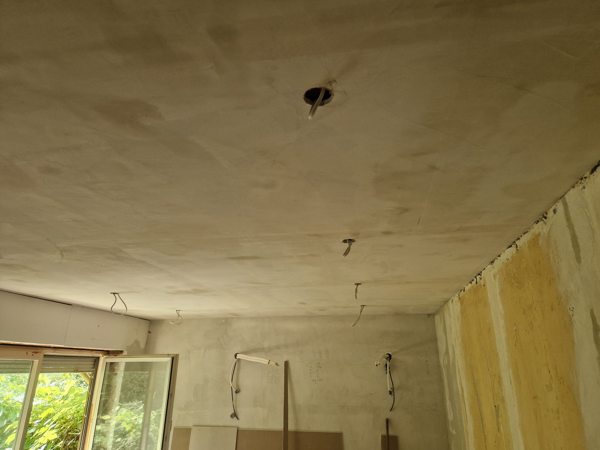 Wnętrze w trakcie prac wykończeniowych: surowy, szpachlowany sufit z wystającymi przewodami elektrycznymi, ściany częściowo z resztkami starej farby, otwarte okno z widokiem na zieleń, płyty...