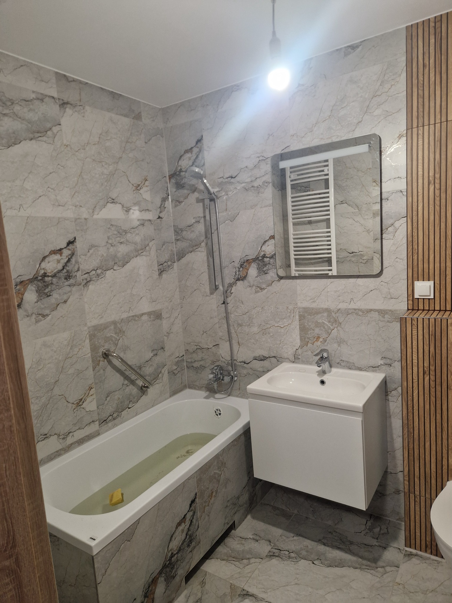 Wykończona łazienka z wanną w zabudowie z płytek marmurkowych, białą szafką z umywalką i lustrem. Na ścianie dekoracyjne panele drewniane i grzejnik drabinkowy.