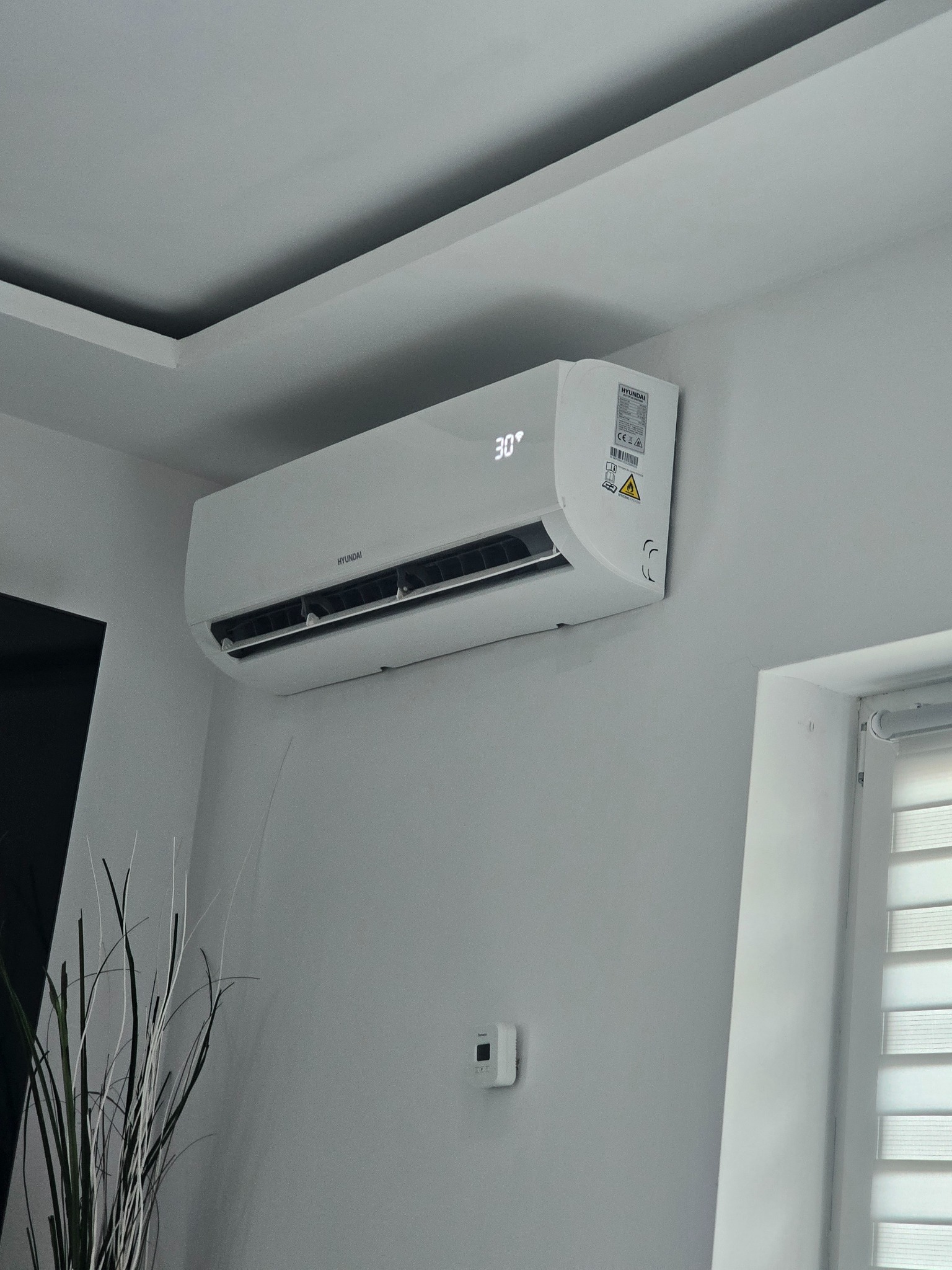 Biały klimatyzator Hyundai zamontowany na ścianie w jasnym pomieszczeniu, wyświetlający temperaturę 30 stopni Celsjusza, obok okno z żaluzjami i dekoracyjna roślina.