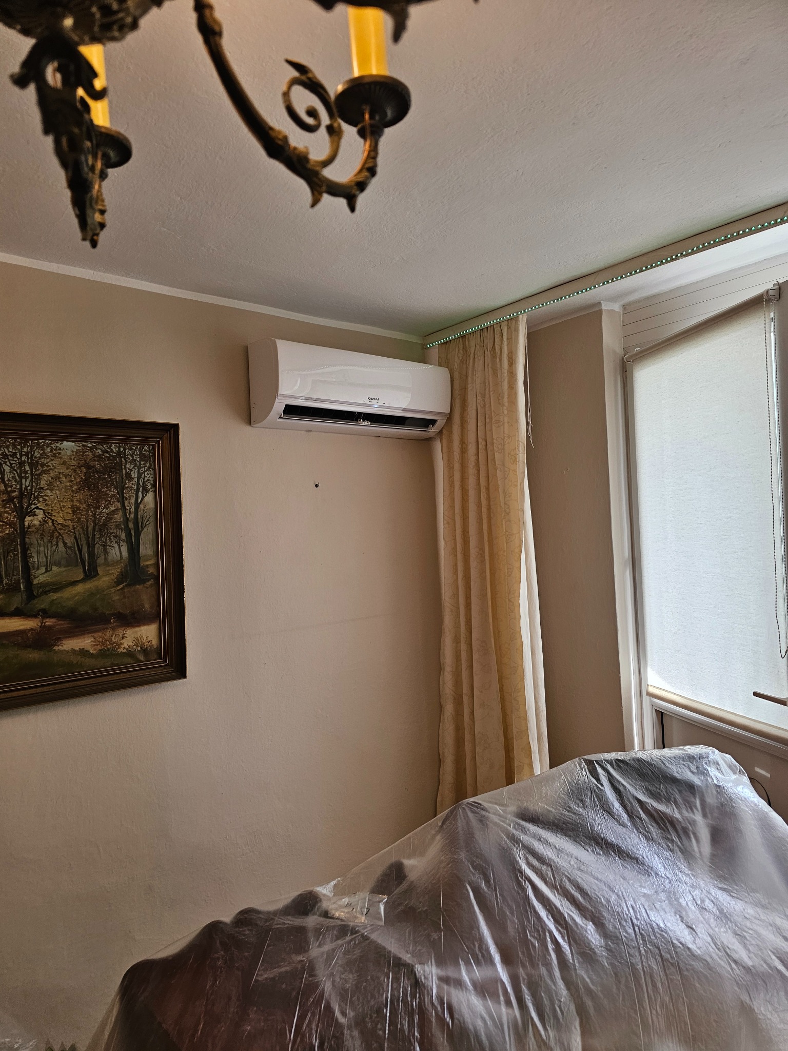 Biała jednostka klimatyzacji zamontowana na ścianie w pokoju z zasłoniętym meblem folią ochronną, obok okna z roletą i obrazu z motywem leśnym; widoczny również żyrandol.