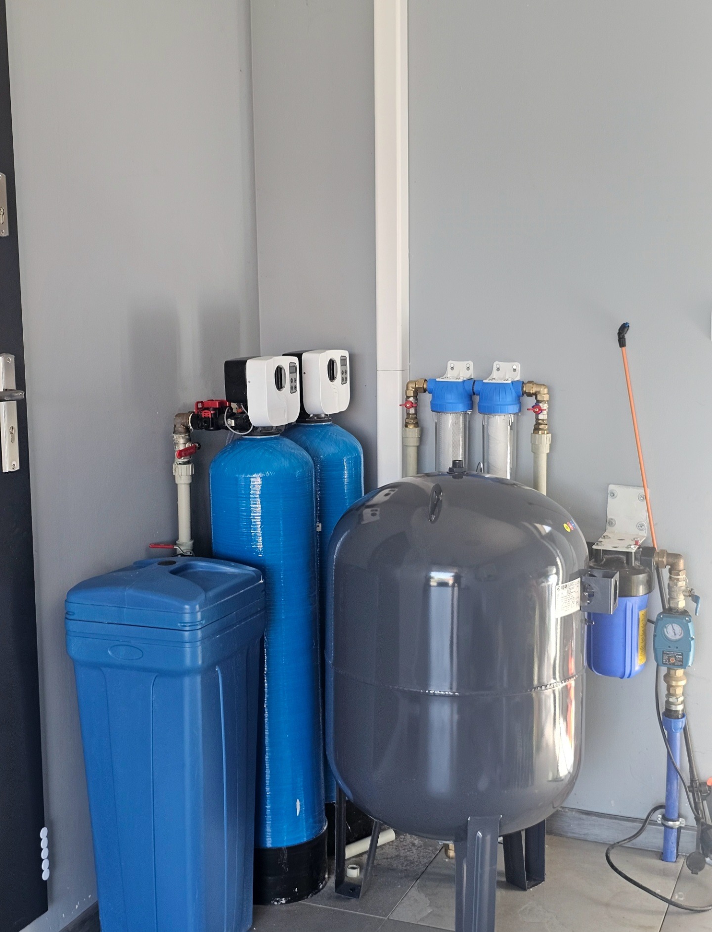 Domowa stacja uzdatniania wody z niebieskimi zbiornikami i szarym zbiornikiem ciśnieniowym, zainstalowana w pomieszczeniu z szarą ścianą i czarnymi drzwiami.