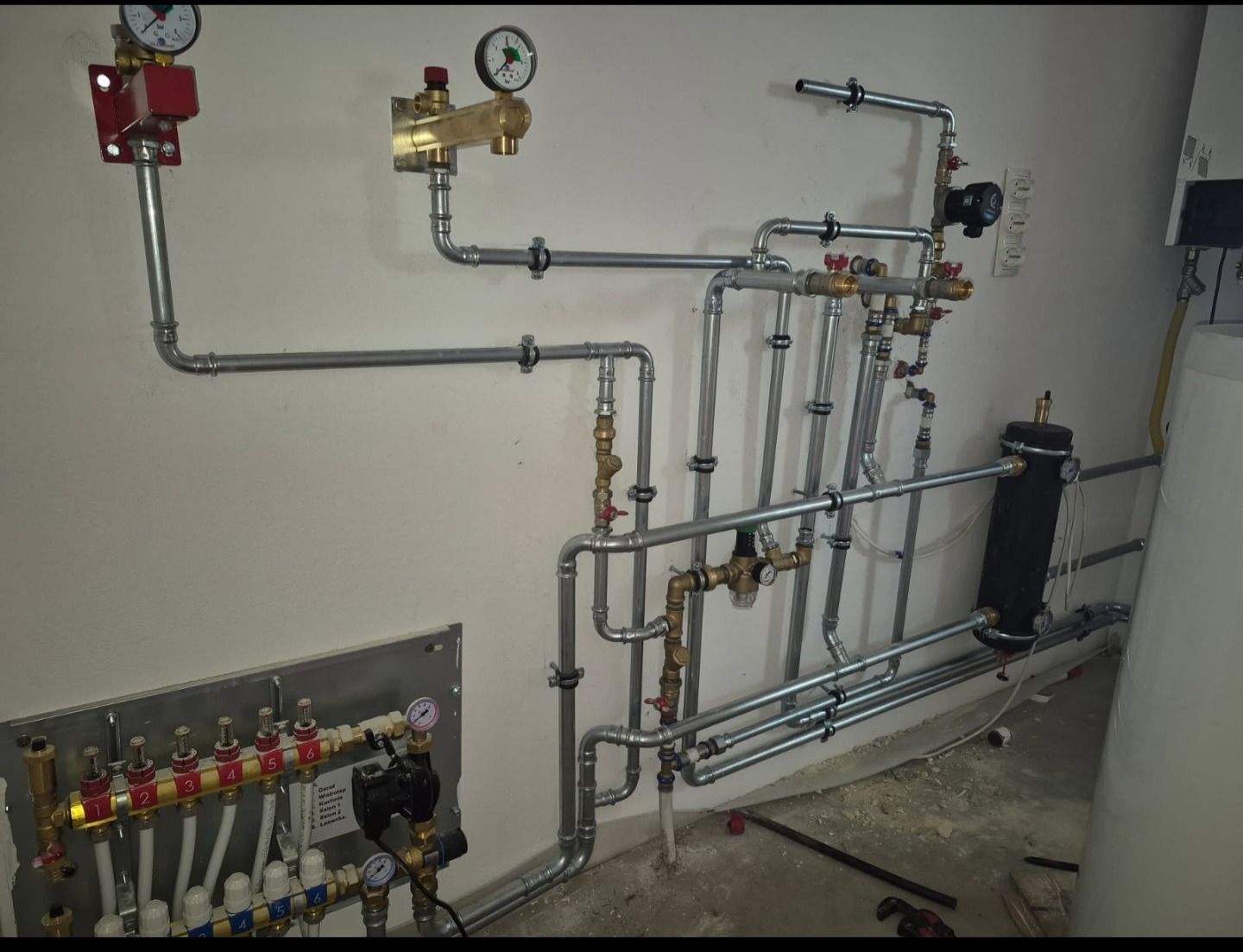 Złożona instalacja hydrauliczna z rur, zaworów i manometrów na ścianie w pomieszczeniu. Widoczny rozdzielacz ogrzewania podłogowego i zbiornik.
