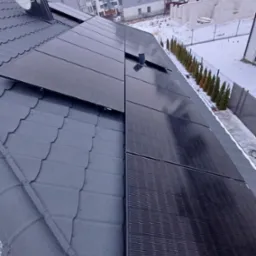 Czarne panele słoneczne na szarym dachu skośnym pokrytym śniegiem, w tle budynki mieszkalne i częściowo zasypany śniegiem teren.