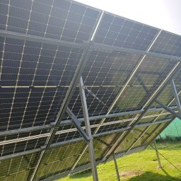 ECOWELL SPÓŁKA Z OGRANICZONĄ ODPOWIEDZIALNOŚCIĄ - Widok od spodu na panele słoneczne zamontowane na metalowej konstrukcji, z widocznym trawnikiem i fragmentem siatki ogrodzeniowej w tle.